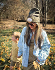 A woman in Faire’s Patch Beanie Support Your Local Mother, sunglasses, and a light denim jacket holds hands with a young boy in tan as they walk outdoors on leaf-covered grass with trees in the background.