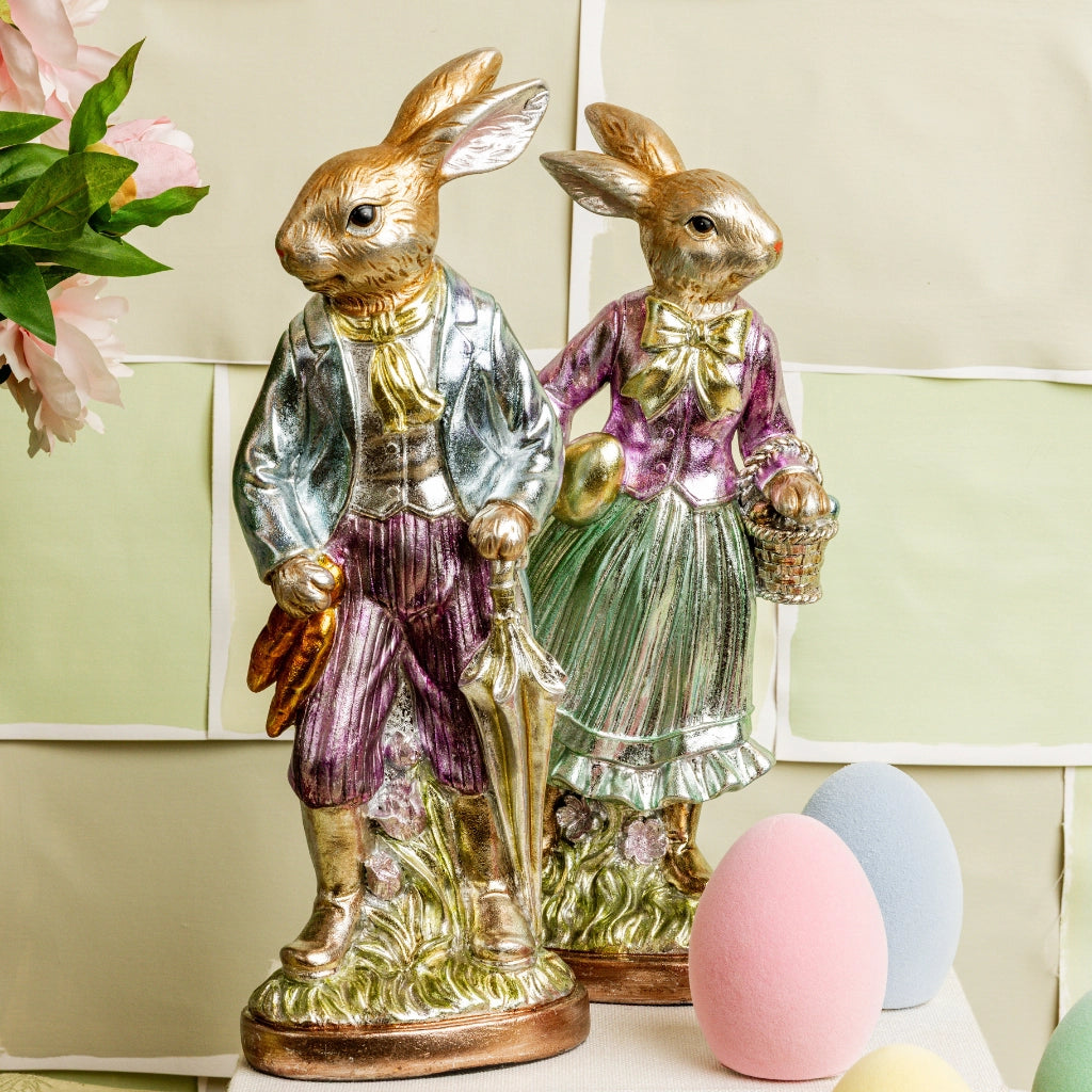 Two Faire Vintage Foil Rabbit figurines stand side by side in heirloom-inspired decor—one with an umbrella, one with a basket—among pastel eggs and pink flowers against a pale checkered background.