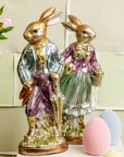 Two Faire Vintage Foil Rabbit figurines stand side by side in heirloom-inspired decor—one with an umbrella, one with a basket—among pastel eggs and pink flowers against a pale checkered background.