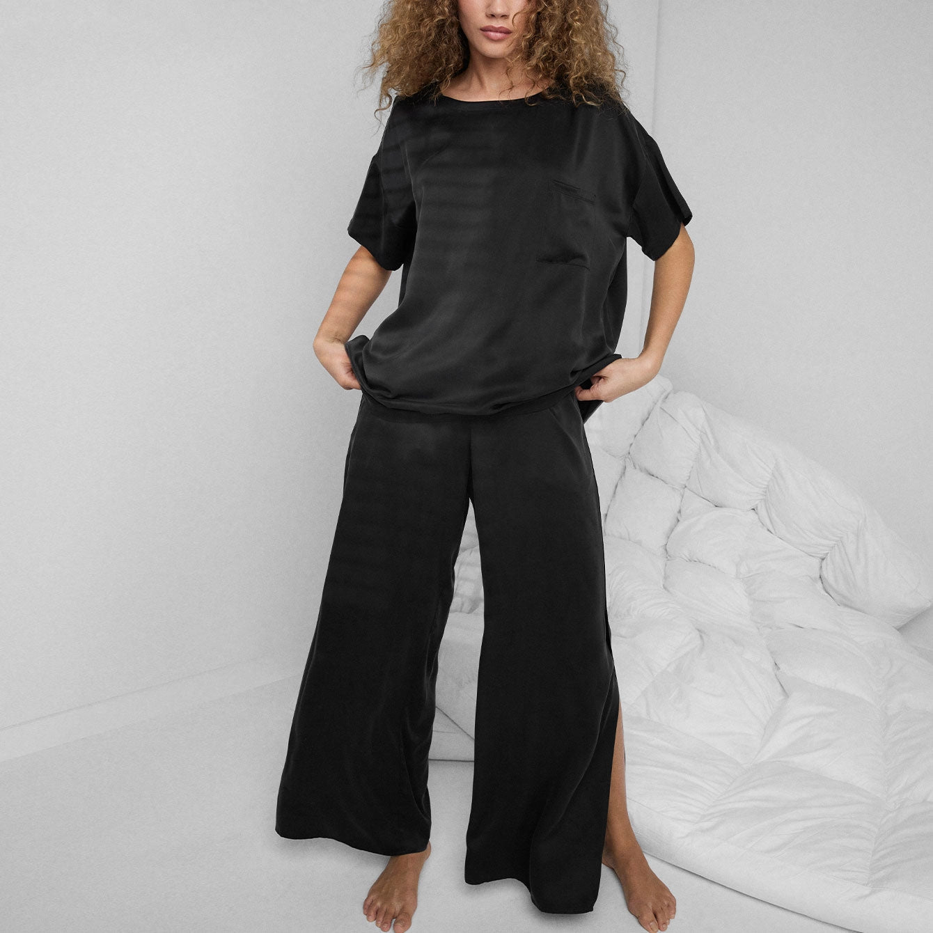 A woman with curly hair stands barefoot in a minimal white room, wearing the Faire Lunya Washable Silk Tee and matching wide-leg pants in black satin. A white, quilted blanket is visible in the background.