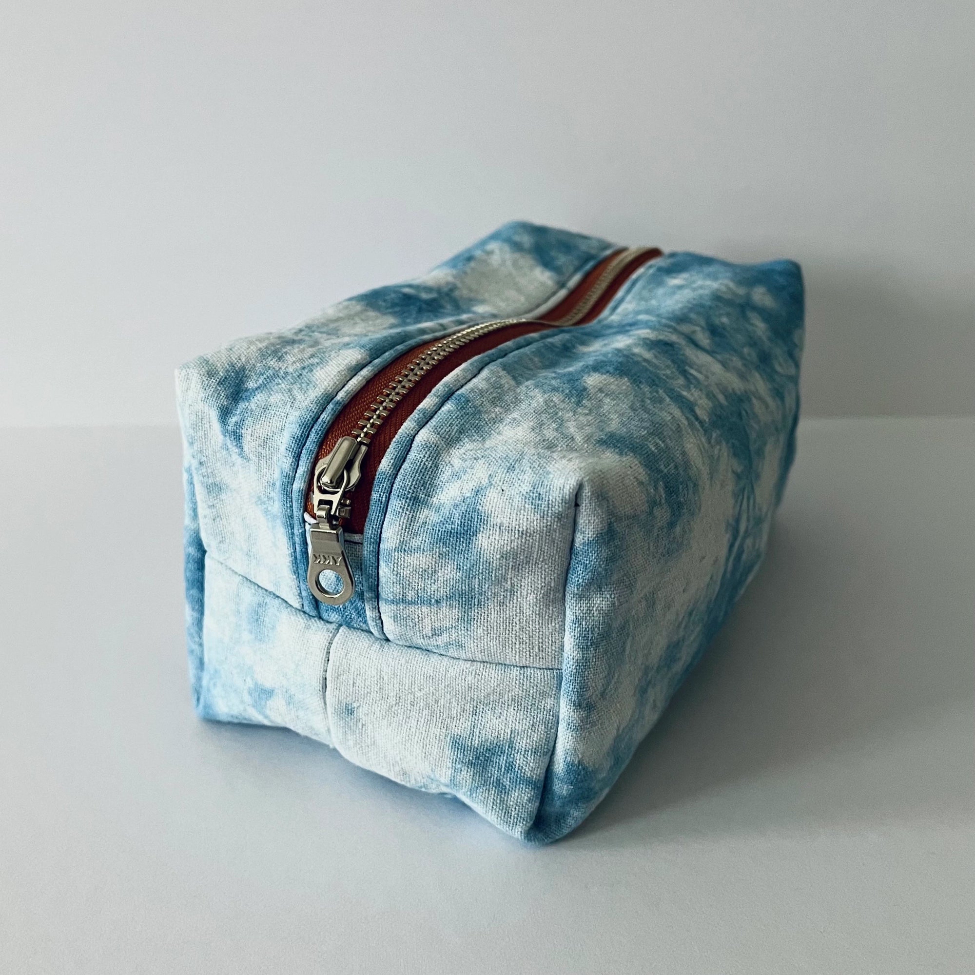 The Faire Dyed Toiletries Bag is a rectangular, hand-dyed blue and white cotton pouch with a metal zipper and red trim, shown on a light surface—ideal for organizing your desk.