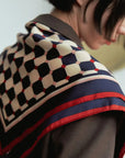 A person with short black hair is wearing the Faire Checkerboard Silk Bandana Americana, which features hand-rolled edges and a geometric pattern in navy, red, and white over a brown outfit. The scarf design includes connected hexagons and dots. The person is facing slightly away from the camera.