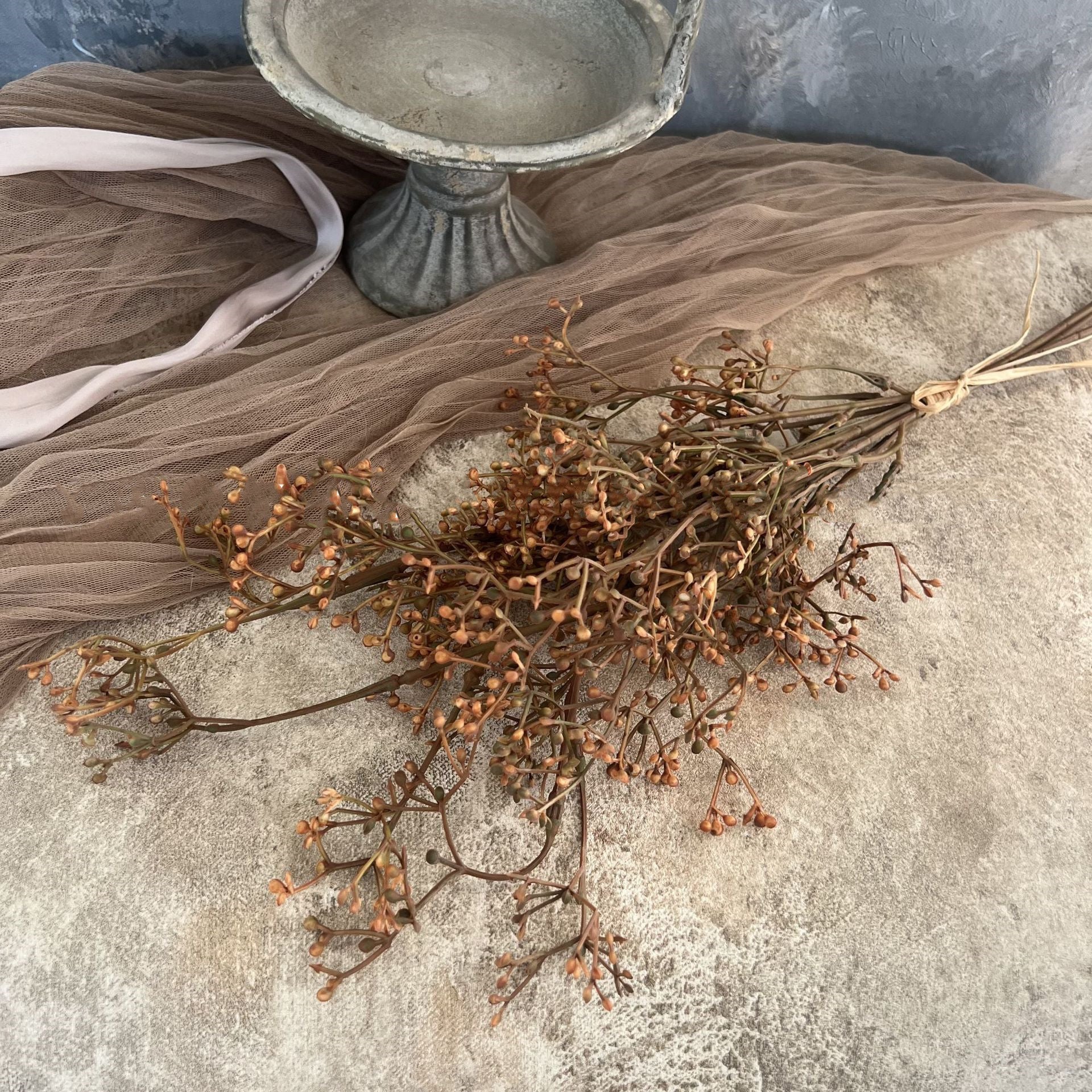 The Faire 22'' Faux Seeded Baby's Breath Bunch is displayed on a textured beige surface next to a gray stone pedestal and sheer taupe fabric, with delicate faux stems adding charm.