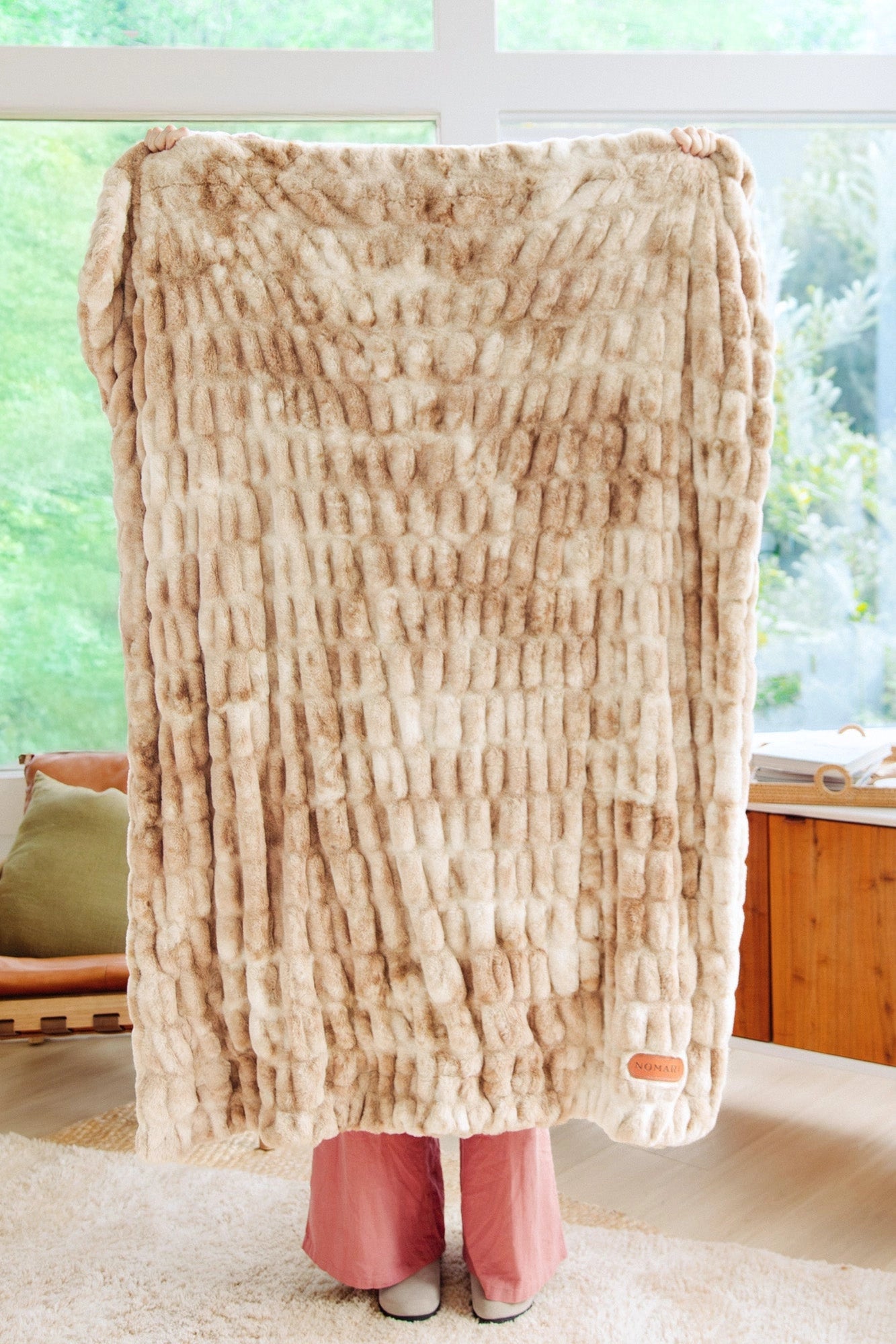 A person indoors holds up the Faire Cascade Throw Blanket, a large beige and white plush faux fur piece that covers their upper body and face. Only their hands and feet show in a room with big windows and wooden furniture.