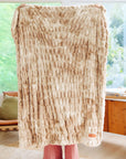 A person indoors holds up the Faire Cascade Throw Blanket, a large beige and white plush faux fur piece that covers their upper body and face. Only their hands and feet show in a room with big windows and wooden furniture.