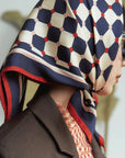 A person drapes the elegant Faire Checkerboard Silk Bandana Americana with hand-rolled edges over their head, complemented by dark sunglasses. The bandana flaunts a chic blue, white, and red geometric design. They pair this look with a brown coat and a patterned inner shirt against an artistically blurred background.