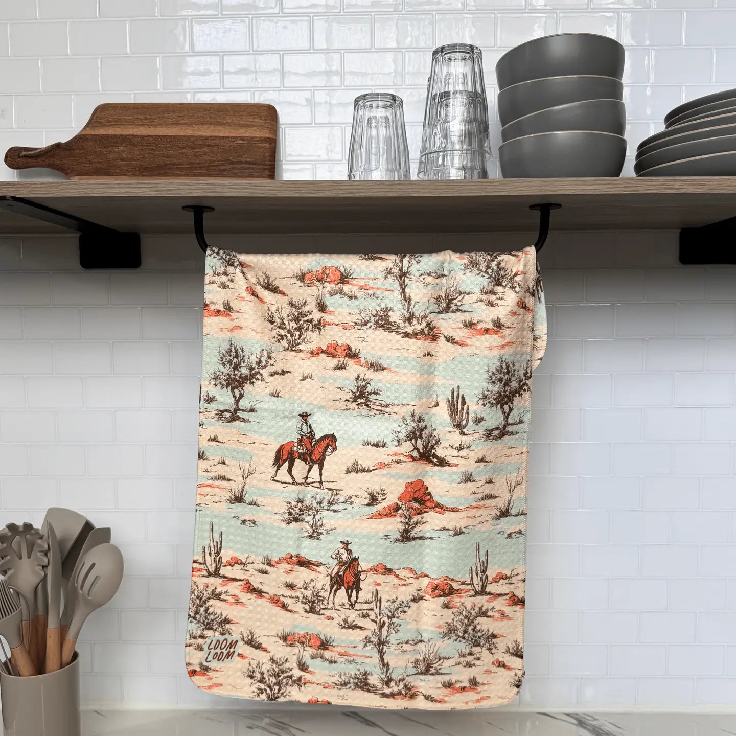 A kitchen displays Faire’s Faded Desert Towel, a super absorbent towel with cowboys on horseback in a desert scene. Plates, bowls, and glasses are stacked above, while cooking utensils are neatly organized below.
