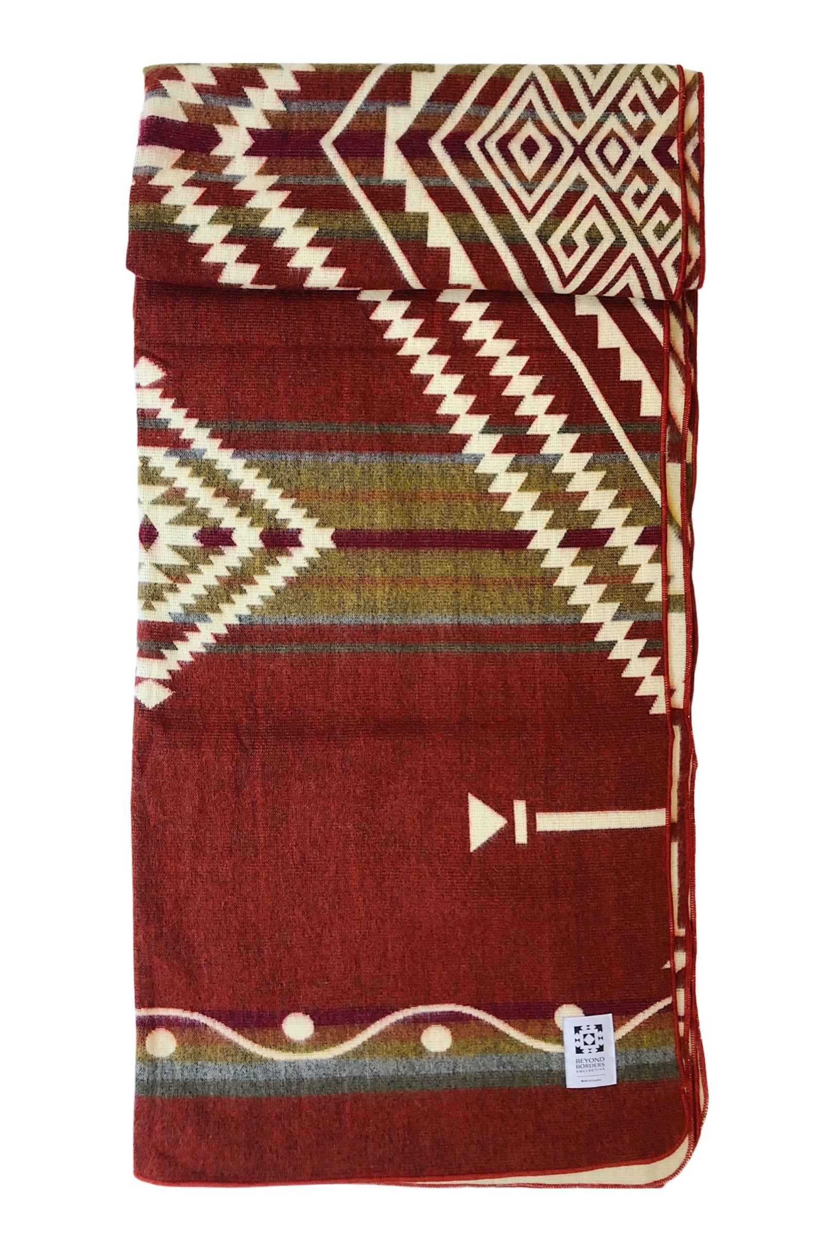 The Achuar Blanket by Faire is a folded Ecuadorian blanket with red, cream, and green geometric patterns—zigzags, diamonds, and arrows—and a small white label in one corner.
