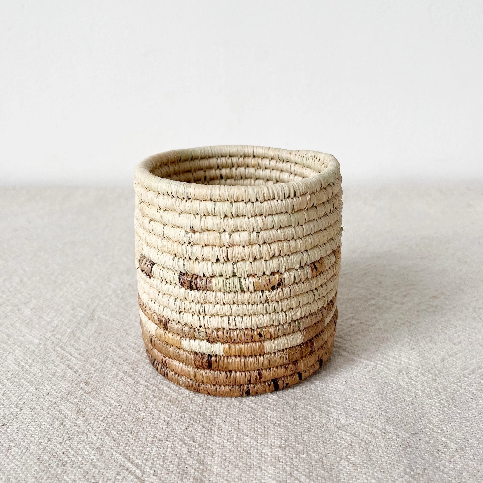 The Mkongo Plant Basket by Amsha - Faire is a small, round, handmade Rwanda basket with beige and brown bands, displayed on a textured, light surface against a plain white background.