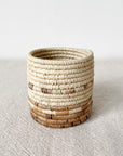 The Mkongo Plant Basket by Amsha - Faire is a small, round, handmade Rwanda basket with beige and brown bands, displayed on a textured, light surface against a plain white background.