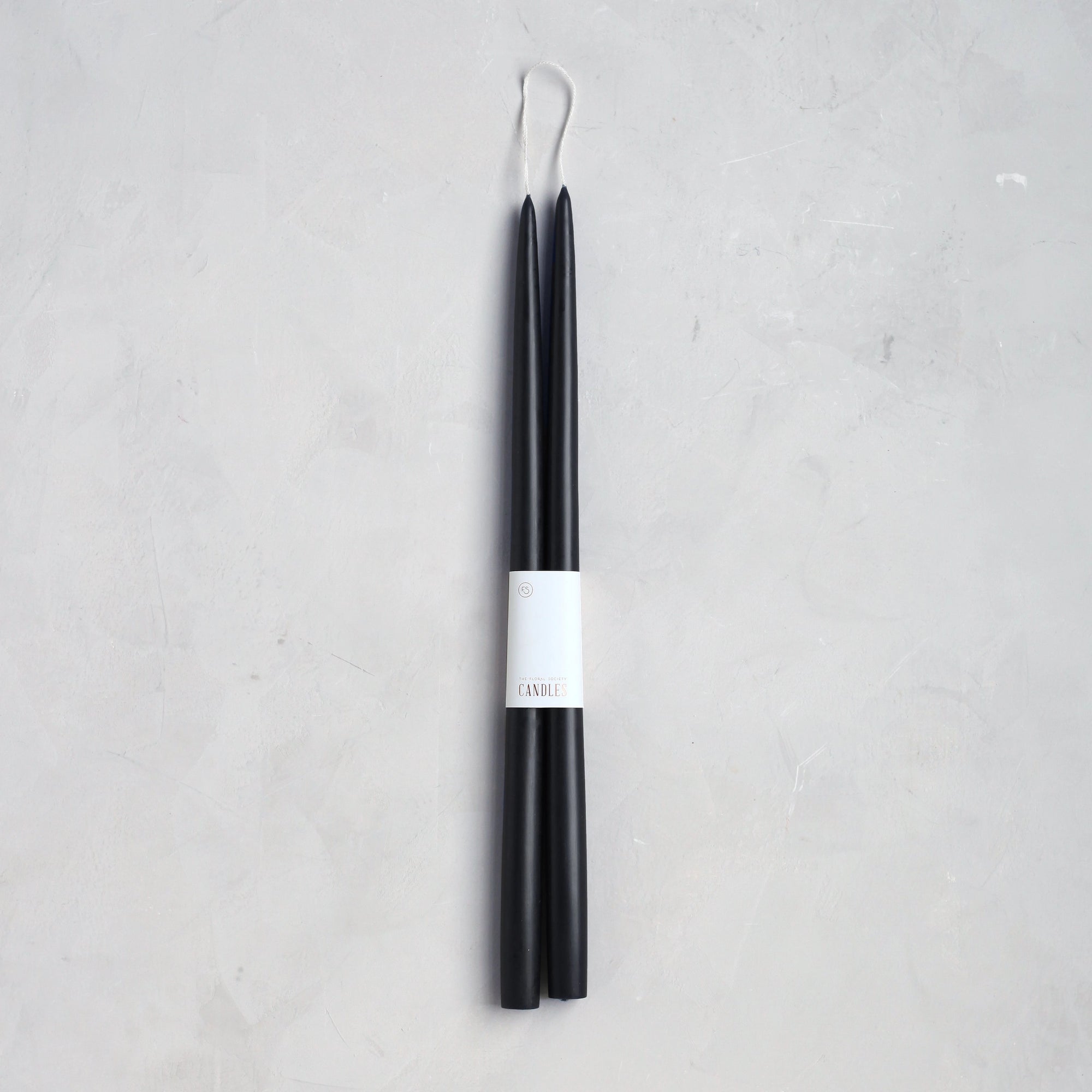 The Floral Society’s 18" Taper Candle - Pair is wrapped in a white paper label, set against a light gray textured background—perfect for adding an elegant touch to any home décor.