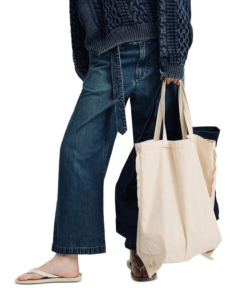 Dressed in Denimist's OVERSIZED ARAN SWEATER W/ROLLED NECK and loose jeans, a person carries a beige tote and dark bag. Casual flip-flops complete this relaxed, thoughtfully accessorized look.