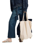 Dressed in Denimist's OVERSIZED ARAN SWEATER W/ROLLED NECK and loose jeans, a person carries a beige tote and dark bag. Casual flip-flops complete this relaxed, thoughtfully accessorized look.