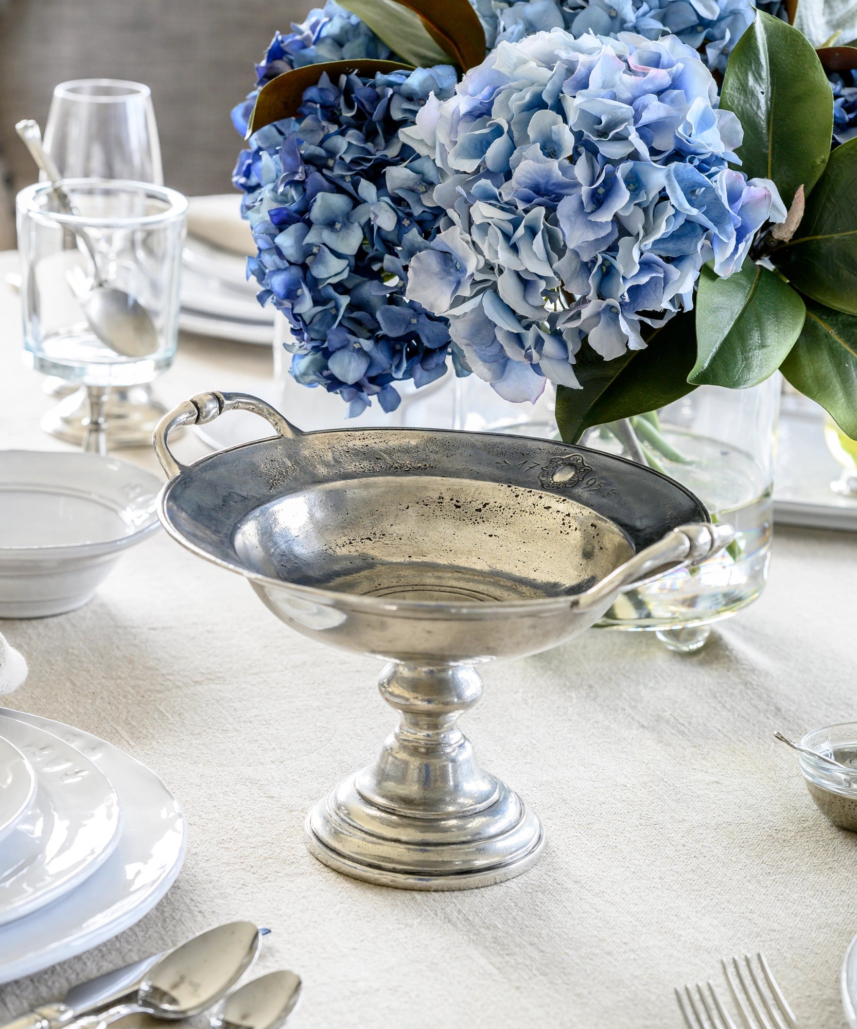 The Vintage 1795 Pedestal Bowl by Faire rests on a cream tablecloth, surrounded by white dishes, silverware, glassware, and a vase filled with blue hydrangeas and green leaves.
