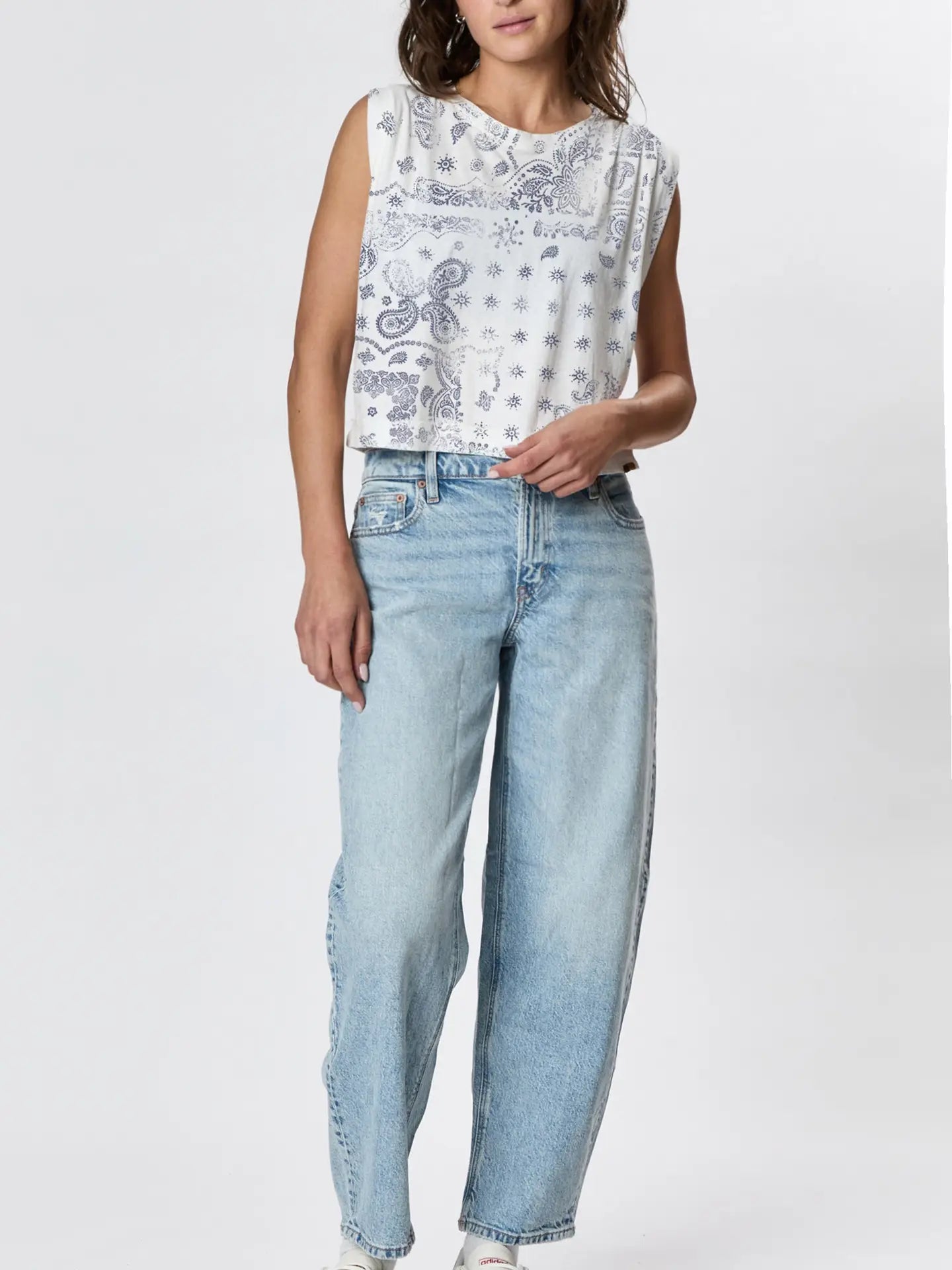 A woman wears the Faire Hemp Jackie Pleat Shell Bandana, a sleeveless white hand-printed shirt with blue paisley and star patterns, paired with light blue high-waisted relaxed-fit jeans, standing against a plain white background.