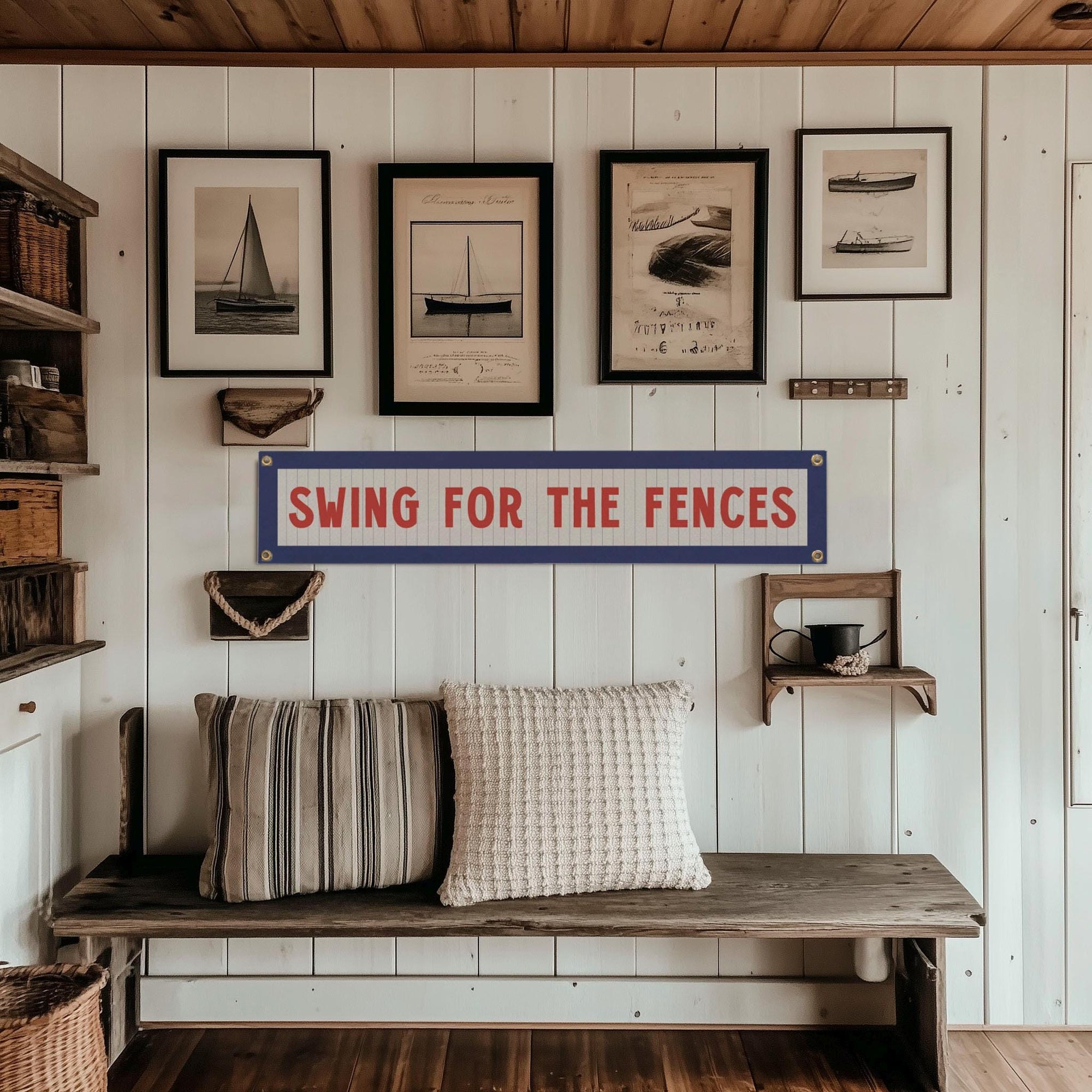 A cozy wooden bench with striped and knitted pillows sits against a white paneled wall. Above, the Faire "Swing for the Fences Banner" adds vintage charm, complemented by neatly arranged framed nautical prints.