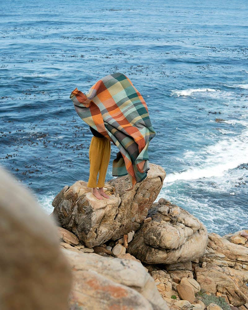 A barefoot person stands on coastal rocks, waves below, mostly wrapped in a Faire Vrou-Vrou Blanket billowing in the wind and obscuring their upper body.