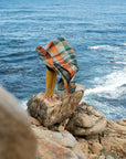 A barefoot person stands on coastal rocks, waves below, mostly wrapped in a Faire Vrou-Vrou Blanket billowing in the wind and obscuring their upper body.