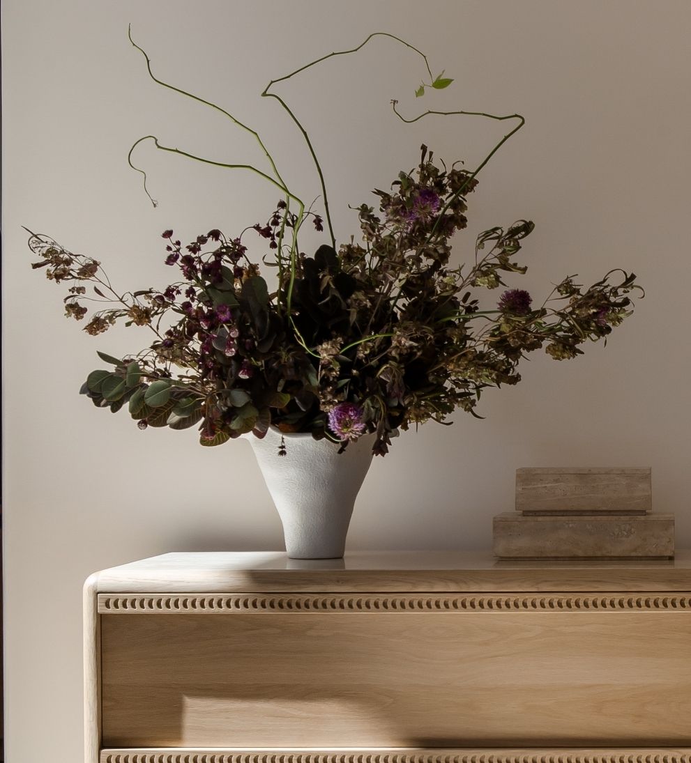 The Andrea Bouquet Vase by Faire, featuring wild dried flowers and greenery, creates a striking centerpiece atop a light wooden cabinet beside two stacked beige books against a softly lit plain wall.