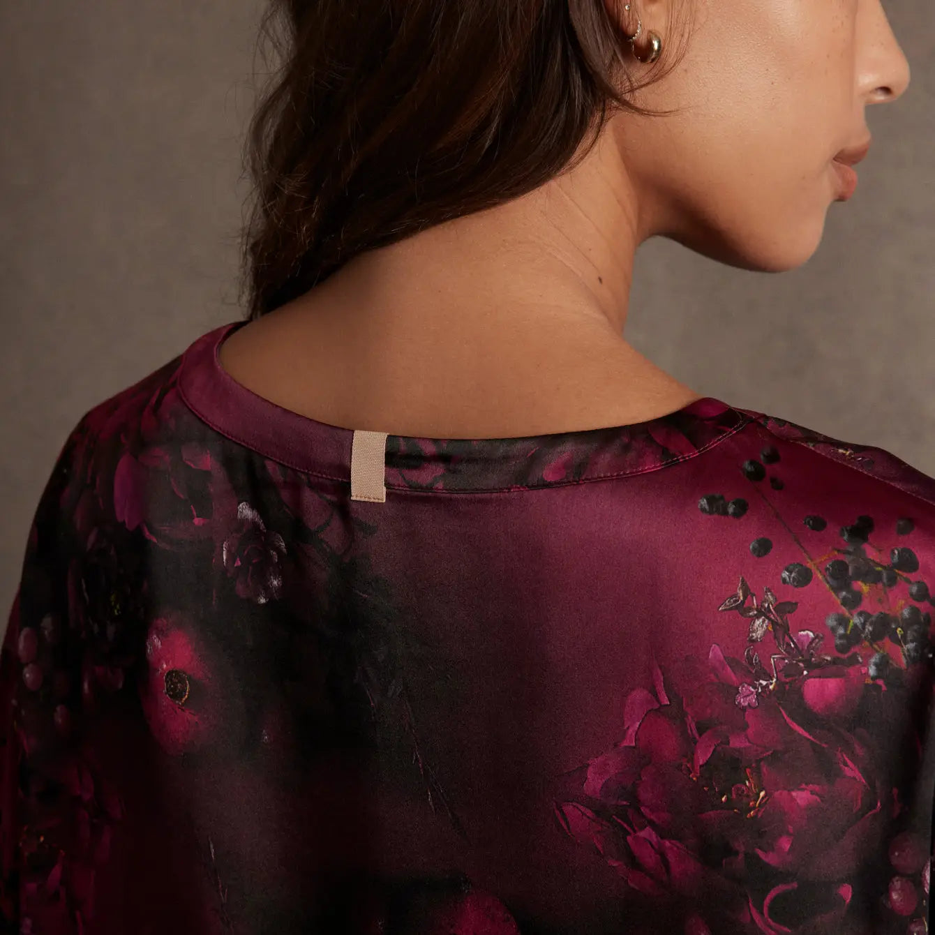 A woman with long brown hair faces away, revealing her bare neck and shoulder in a deep burgundy Lunya Washable Silk Tee by Faire, featuring a floral pattern and small beige neckline tag against a softly lit neutral background.