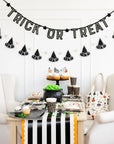 A Halloween table setup features black and white decor, cupcakes, party hats, a cauldron with green candy, a striped table runner, and the My Mind's Eye Bunting Banner glowing above.