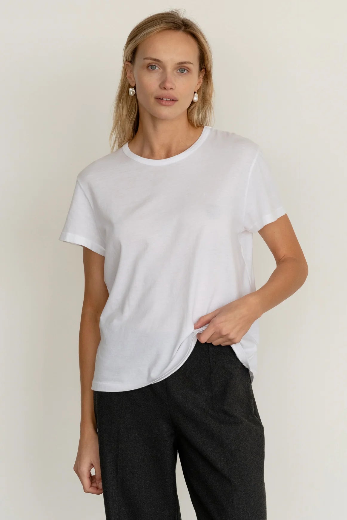A woman with light hair wears Donni’s The Jersey Tee in plain white Supima cotton and dark pants, standing against a neutral background with a relaxed expression and one hand partly in her pocket.