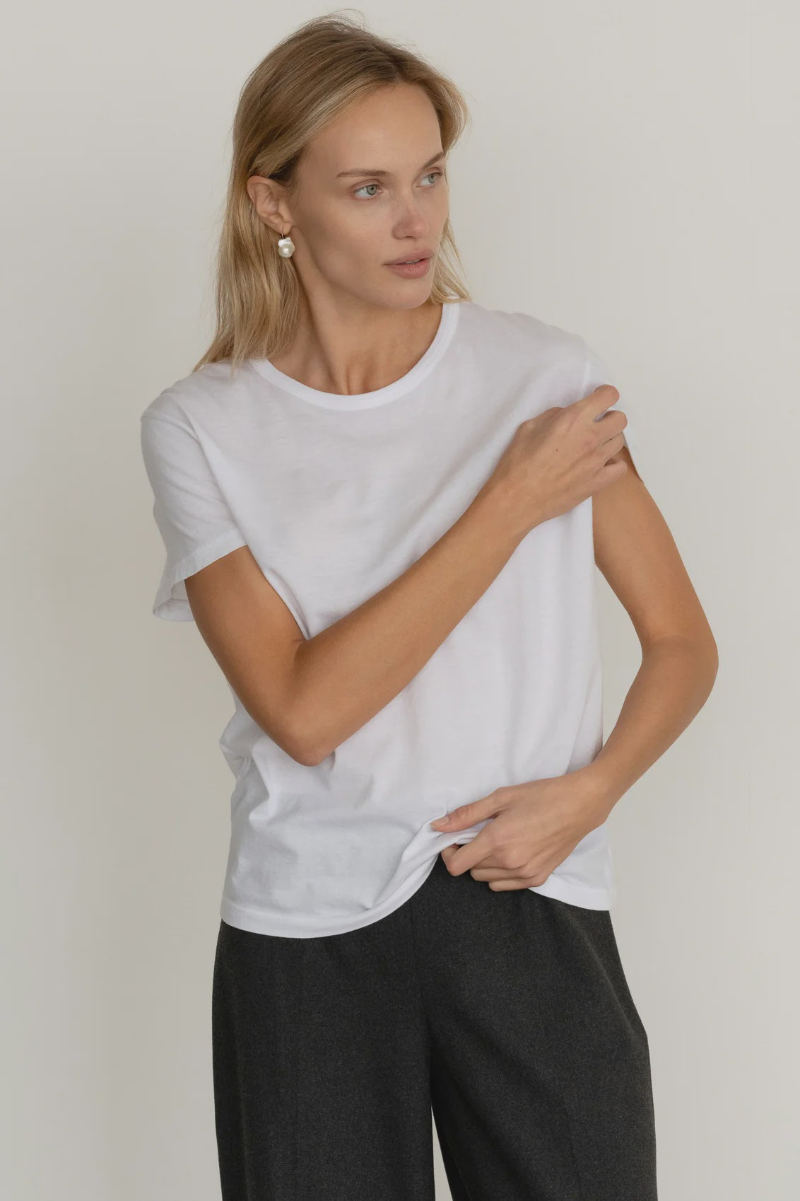 A blonde woman stands against a light background, looking to the side as she adjusts her sleeve, wearing Donni's The Jersey Tee and dark trousers.