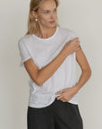 A blonde woman stands against a light background, looking to the side as she adjusts her sleeve, wearing Donni's The Jersey Tee and dark trousers.