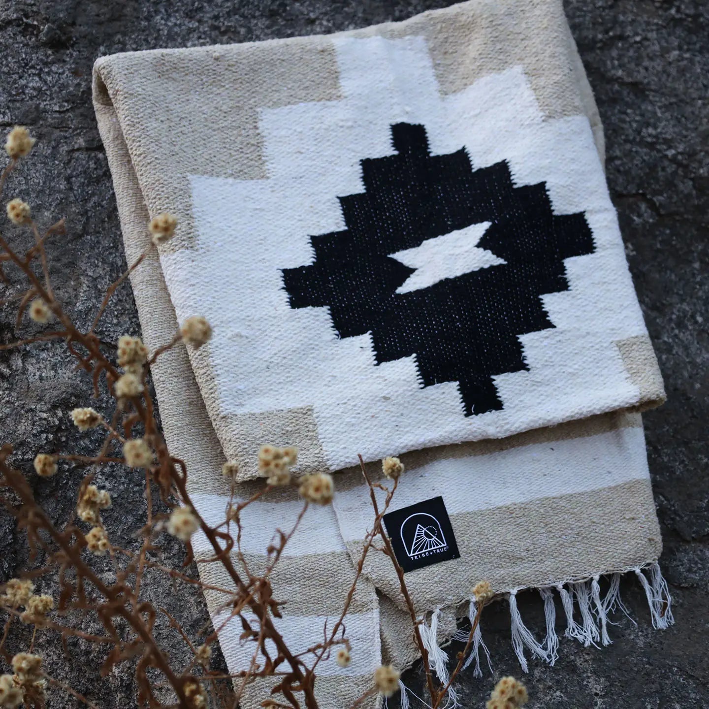 The TT Handwoven Blanket by Faire, featuring a bold black geometric pattern on white and beige, is folded on a rock with dried plants nearby and a small black Faire logo label on the corner.