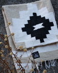 The TT Handwoven Blanket by Faire, featuring a bold black geometric pattern on white and beige, is folded on a rock with dried plants nearby and a small black Faire logo label on the corner.