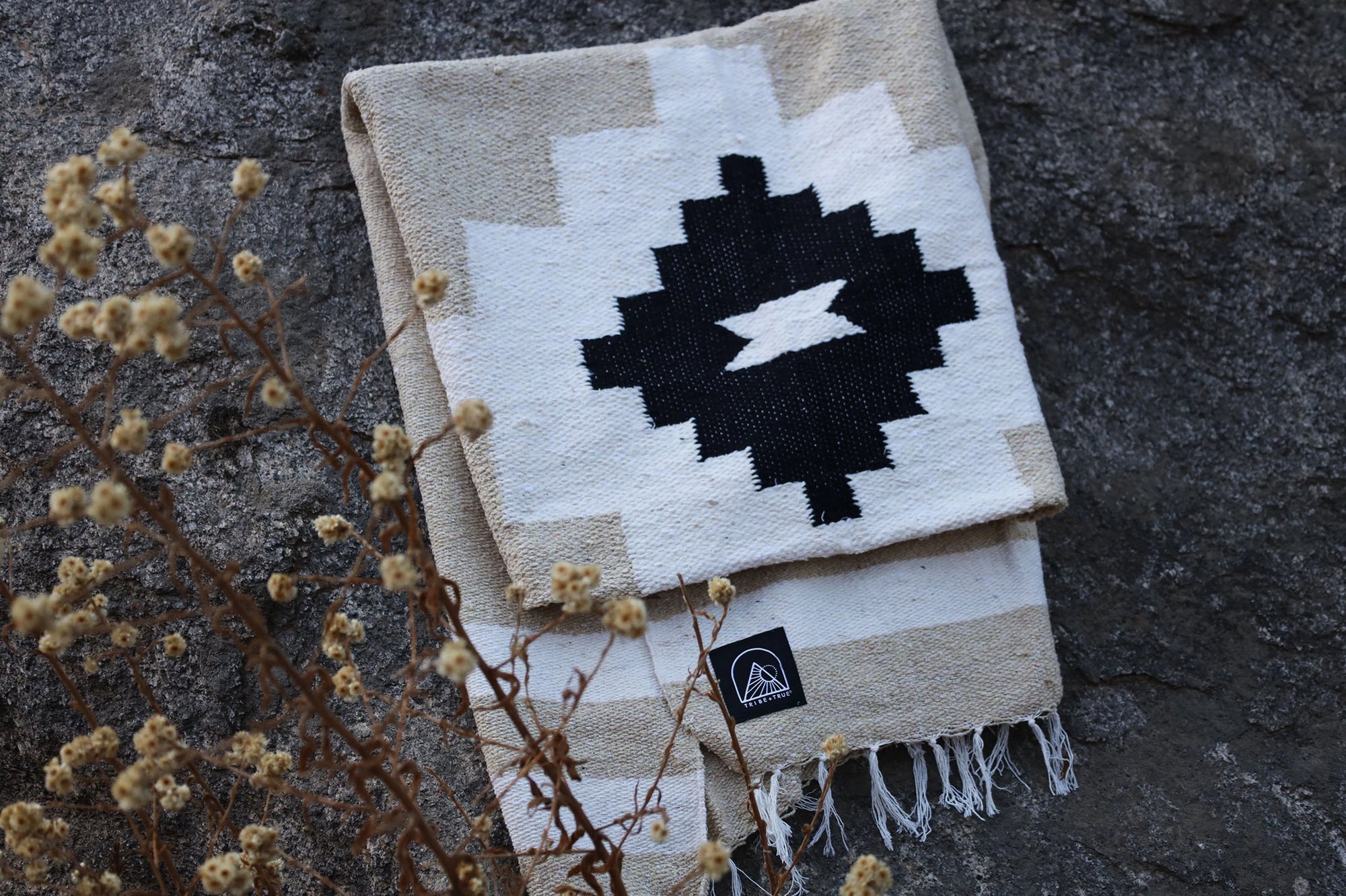 The TT Handwoven Blanket Taos Two by Faire, featuring a bold black geometric pattern on white, is folded on a rough gray stone surface with dried wildflowers in the foreground.