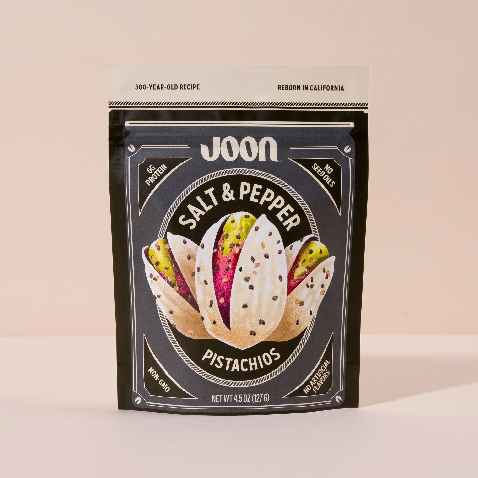 A black and beige resealable pouch of Faire Joon Pistachios stands upright. The front shows pistachio illustrations and highlights this healthy snack as high protein, non-GMO, and with no added sugar.