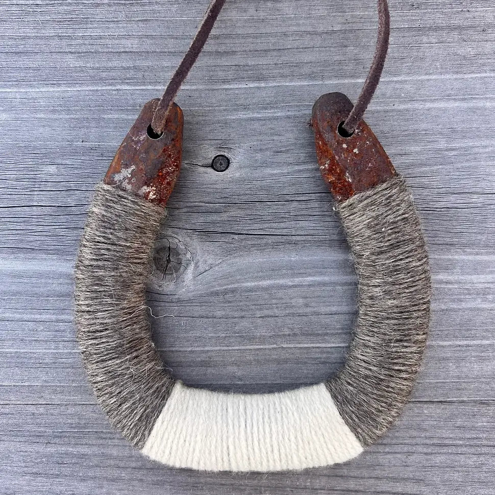 The Faire Lucky Horseshoe features upcycled metal wrapped in gray and white yarn, hanging by a leather cord against a weathered wood background—a perfect rustic decor piece.