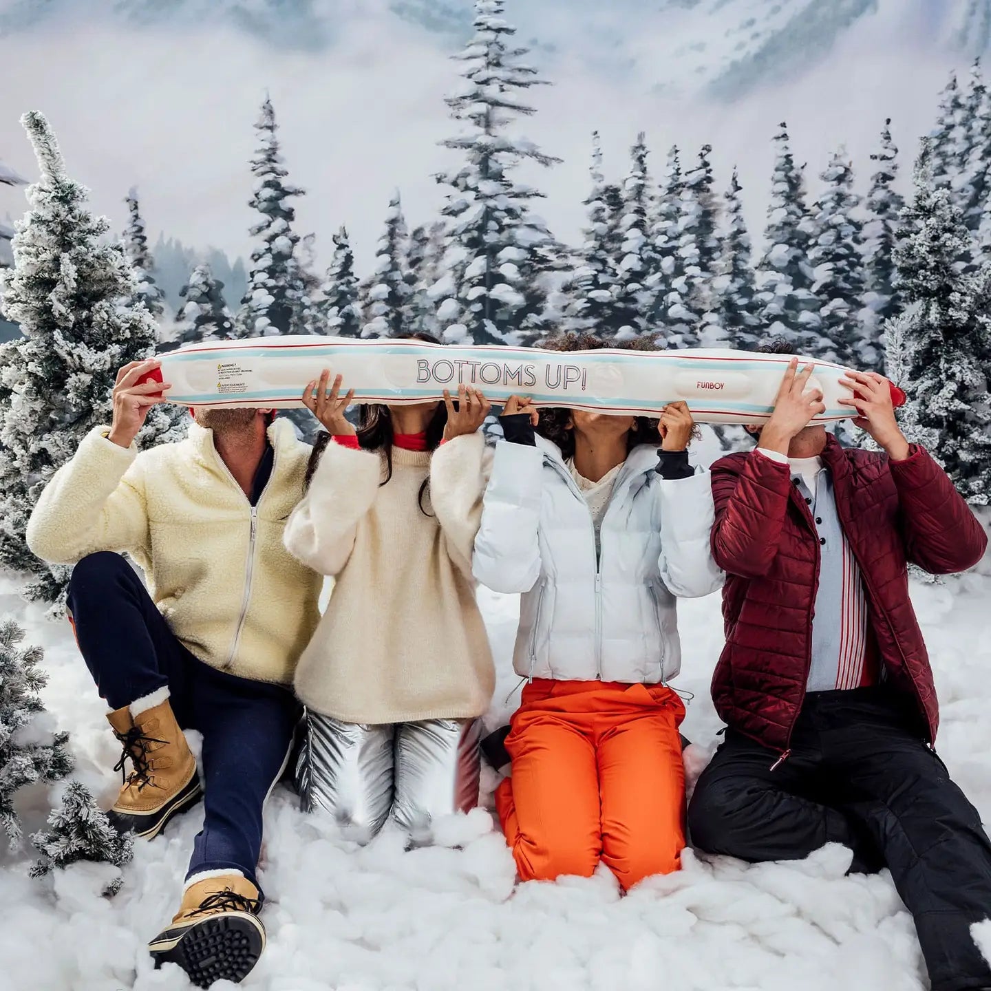 Four people in winter clothing are gathered outside in a snowy landscape, enjoying their drinks with the inflatable Apres Ski Shotski from Faire, labeled "BOTTOMS UP!" All around them are snow-covered trees and majestic mountains.