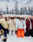 Four people in winter clothing are gathered outside in a snowy landscape, enjoying their drinks with the inflatable Apres Ski Shotski from Faire, labeled "BOTTOMS UP!" All around them are snow-covered trees and majestic mountains.