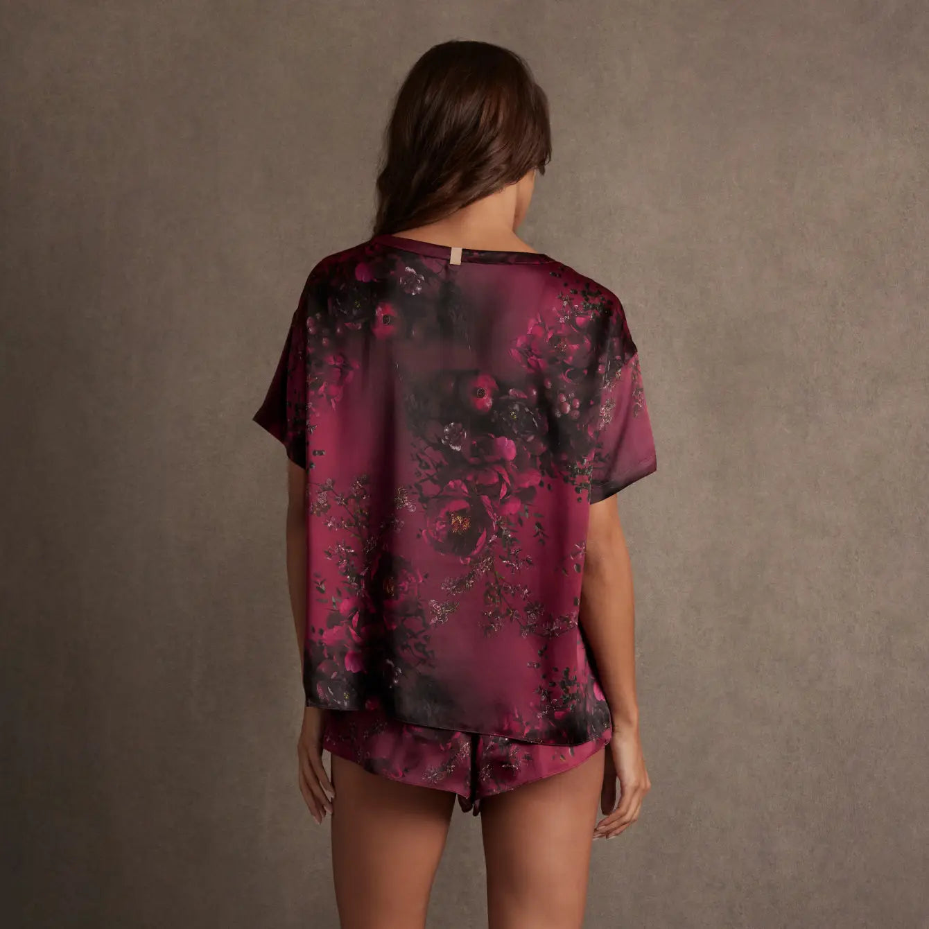 A woman faces away from the camera, wearing the Faire Lunya Washable Silk Tee and matching shorts in a dark floral print. The background is plain and neutral-toned.