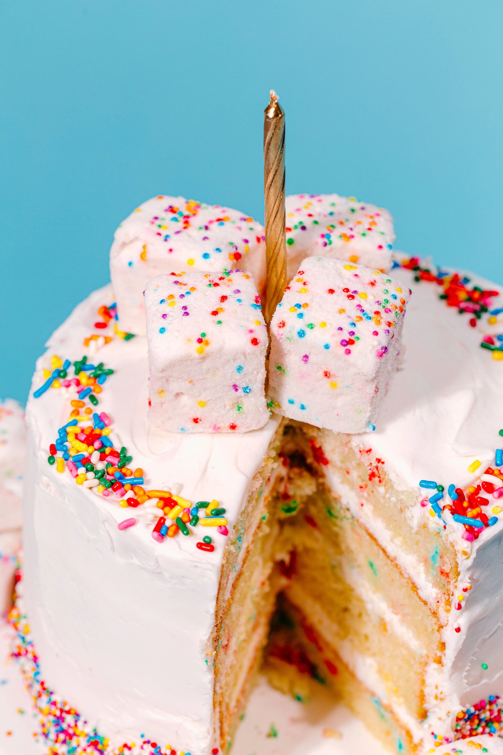 A white frosted layer cake topped with Faire Funfetti Marshmallows, colorful sprinkles, and a gold candle. A slice is cut out to reveal the yellow inside. The backdrop is a solid light blue.