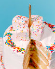 A white frosted layer cake topped with Faire Funfetti Marshmallows, colorful sprinkles, and a gold candle. A slice is cut out to reveal the yellow inside. The backdrop is a solid light blue.