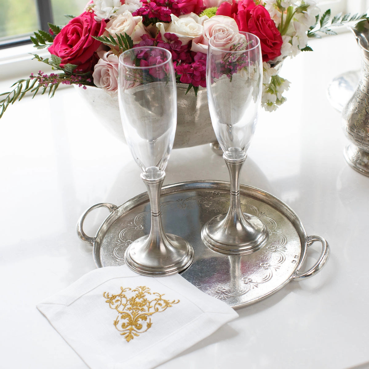 Two empty champagne flutes sit on the Vintage Pewter Round Tray With Handles by Faire, atop a white napkin with gold detail. In the background, an Italian pewter bowl overflows with pink and white flowers.