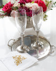 Two empty champagne flutes sit on the Vintage Pewter Round Tray With Handles by Faire, atop a white napkin with gold detail. In the background, an Italian pewter bowl overflows with pink and white flowers.