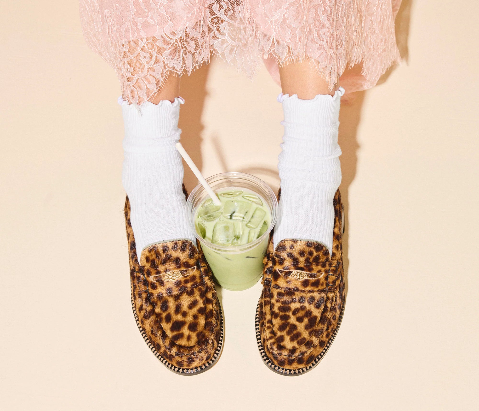 A person wearing a pink lace skirt, white ruffled socks, and the Freda Salvador ELBA Leopard Loafer with a glass of iced green drink with a straw placed between their feet on a light background.