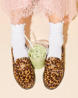 A person wearing a pink lace skirt, white ruffled socks, and the Freda Salvador ELBA Leopard Loafer with a glass of iced green drink with a straw placed between their feet on a light background.