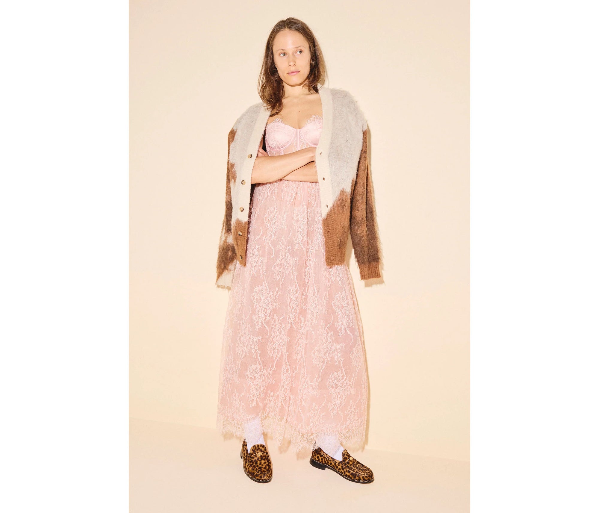 A woman with shoulder-length hair and a neutral expression stands against a beige background, wearing a long pink lace dress, an oversized brown and beige cardigan, white socks, and the Freda Salvador ELBA Leopard Loafers.