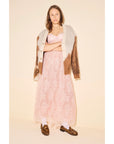 A woman with shoulder-length hair and a neutral expression stands against a beige background, wearing a long pink lace dress, an oversized brown and beige cardigan, white socks, and the Freda Salvador ELBA Leopard Loafers.