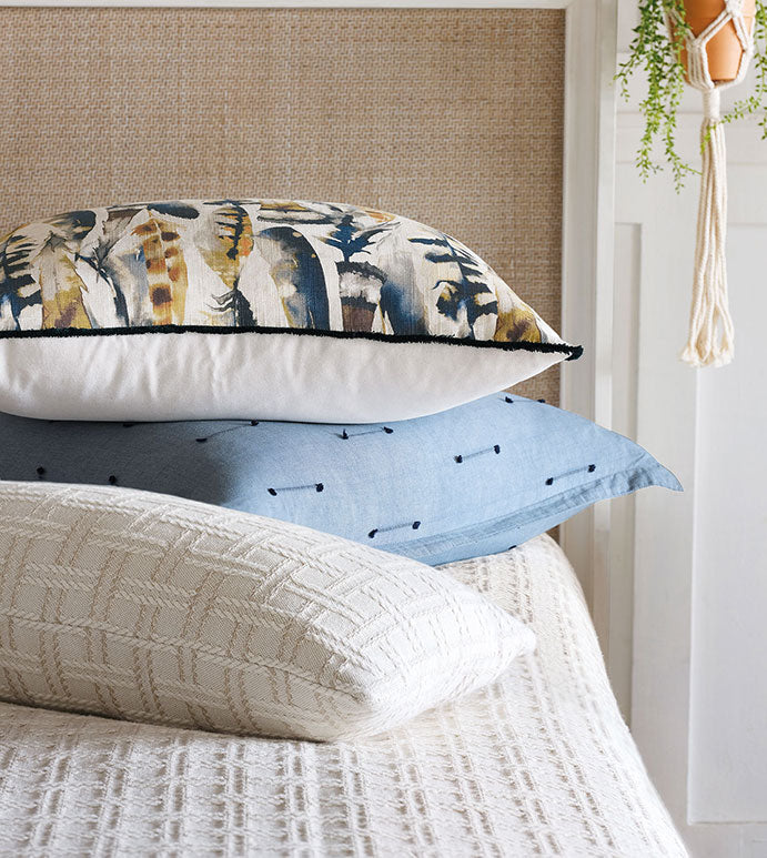 A neatly stacked arrangement of three pillows on a bed. The top pillow, adorned with Eastern Accents&#39; EUS-429 Sprouse Watercolor featuring an abstract feather pattern in various colors, resembles a watercolor print. The middle pillow is a blue cushion with dark blue dots, and the bottom pillow is an off-white, woven-texture cushion. A small hanging plant is visible in the background.