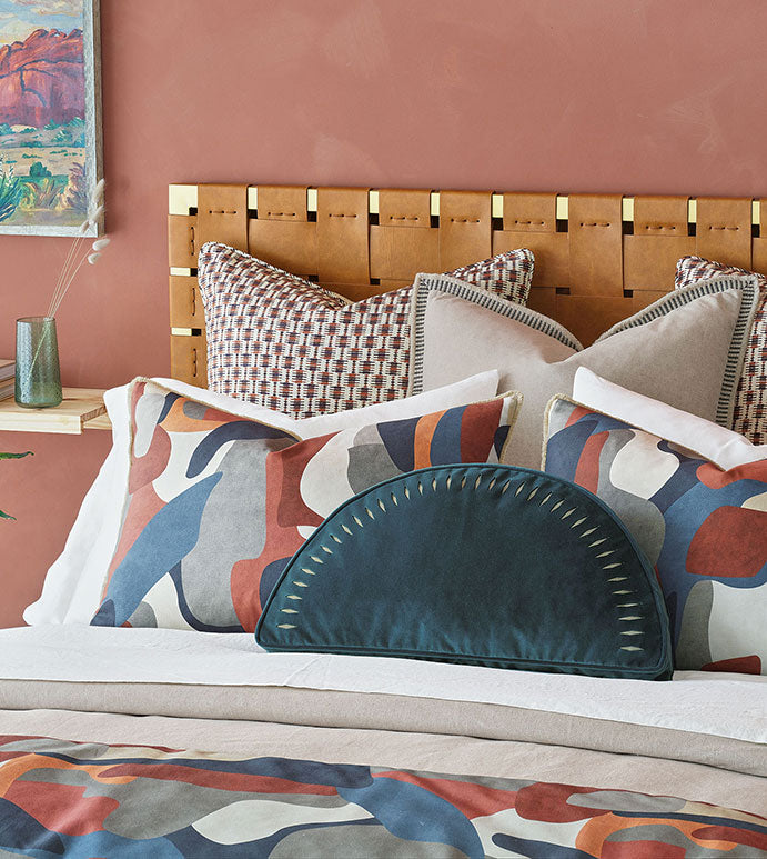 A stylishly decorated bedroom corner in a Scottsdale bungalow featuring a Moab Graphic 27x27&quot; bedspread from Eastern Accents with an artistic headboard against a soft coral wall, adorned with decorative pillows in various patterns and a bedspread with abstract design.