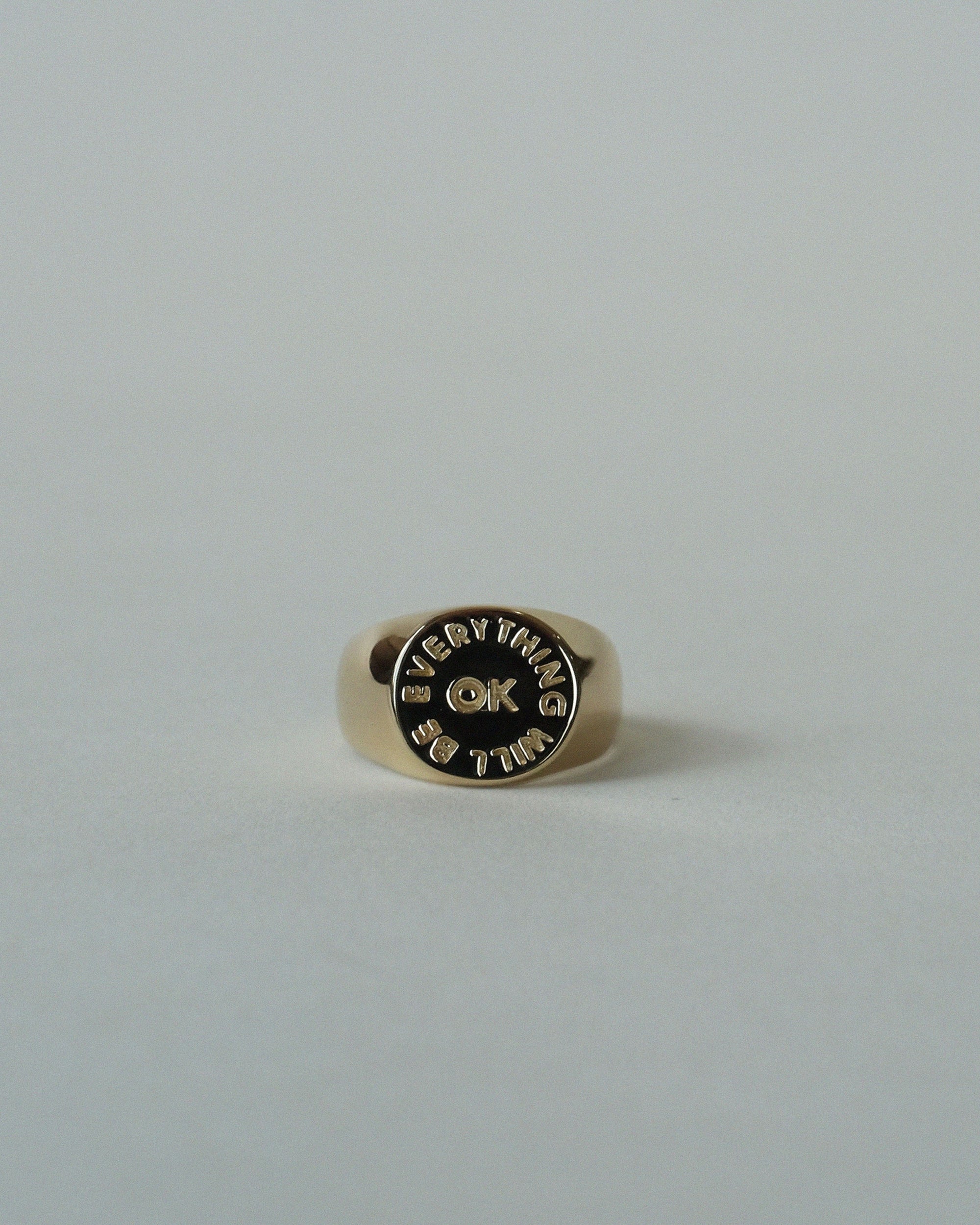 The Faire Everything Will Be OK Ring is a gold-plated signet with “EVERYTHING IS OK” engraved on its round face, shown on a plain light grey background.