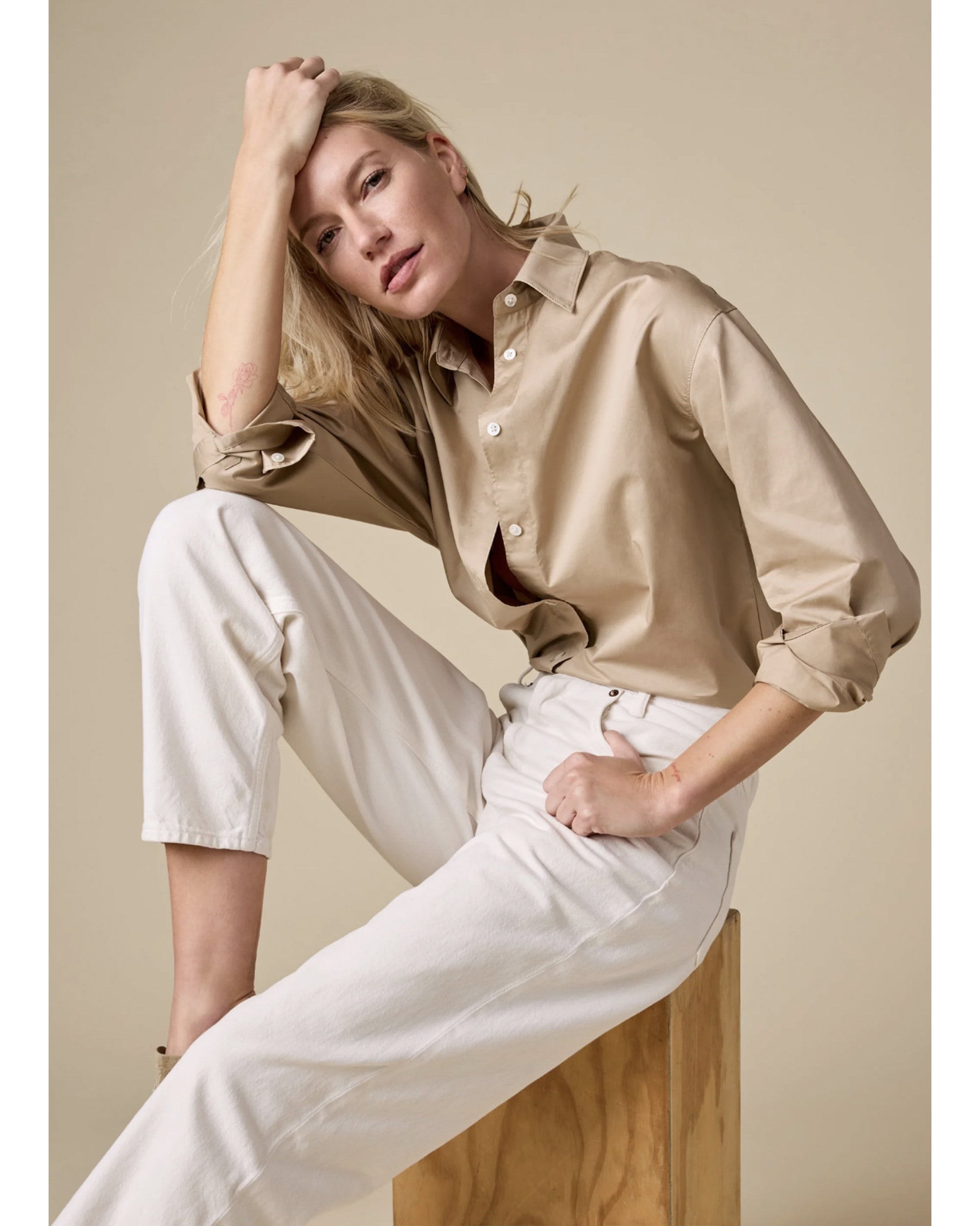 A woman with blonde hair sits on a wooden block against a beige background, wearing The Everything Shirt by Sold Out NYC—an oversized 100% cotton button-up—paired with white pants. She gazes at the camera, resting her elbow on her knee and hand on her head.