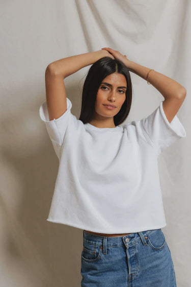 A woman with long dark hair wears the Perfectwhitetee French Terry SS Loose Sweatshirt and blue jeans, standing before a light fabric backdrop with her arms raised above her head.