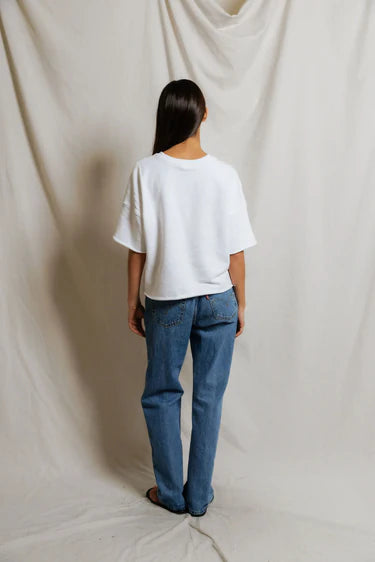 A person with long dark hair, wearing the Perfectwhitetee French Terry SS Loose Sweatshirt in white and blue jeans, stands facing away from the camera against a neutral draped fabric background.